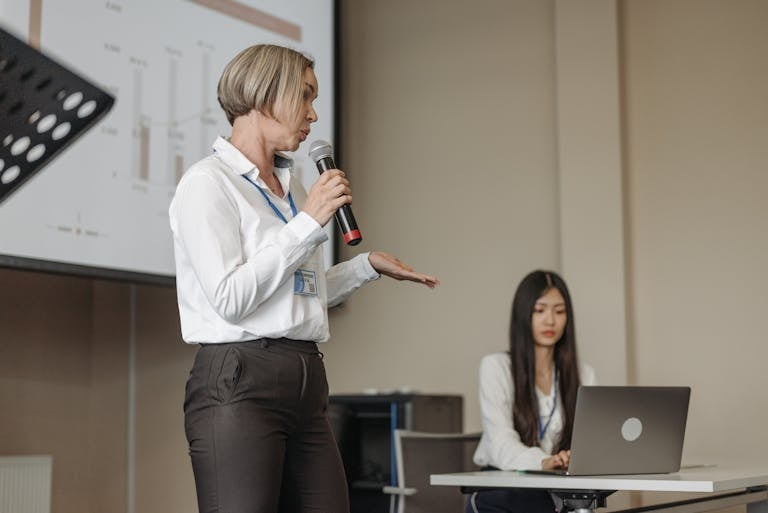 Female speaker presenting at a business conference with an audience member working on a laptop.